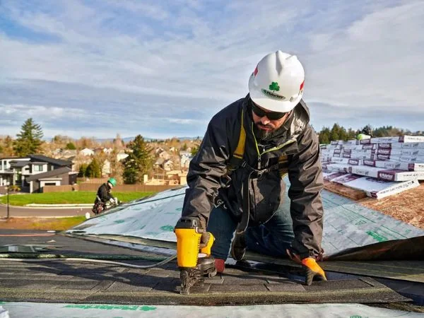 A roofer in a safety harness fastening shingles on a residential roof