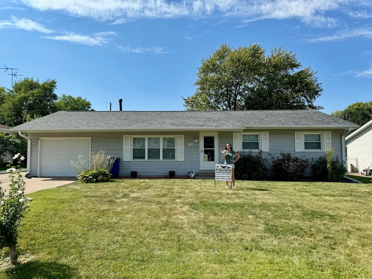 A ranch home with a newly installed shingle roof, the homeowner standing by a Fisher Construction yard sign